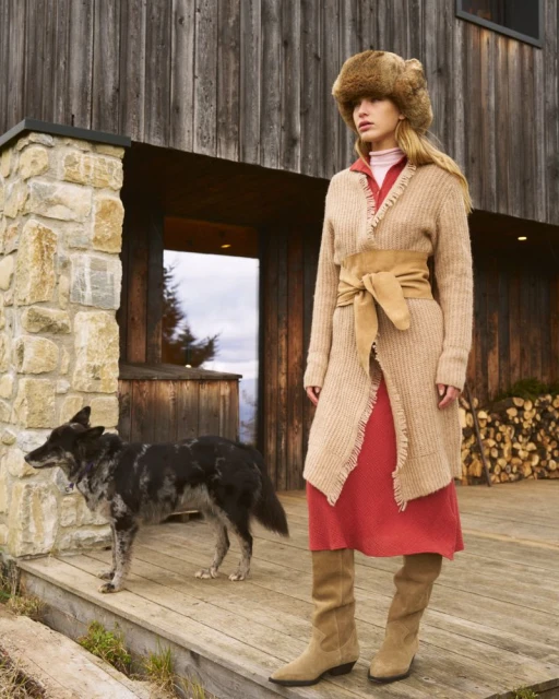 Fashionable woman in fur hat and cardigan with dog poses on wooden porch in front of cabin-style home on cloudy day.