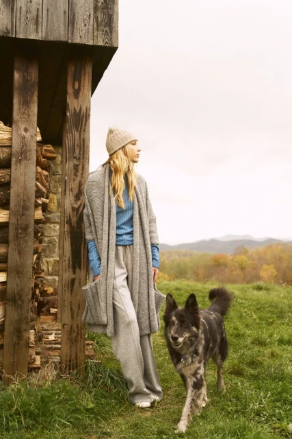 Woman in hat and grey outfit stands next to wood pile in rural setting, accompanied by dog, under an overcast sky. Fashion editorial.