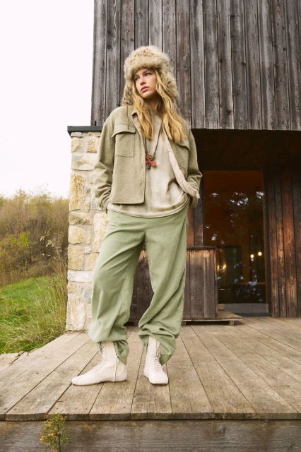 A woman models outdoors on a wooden deck wearing a fur hat, jacket, t-shirt, light green pants, and boots with a rustic building behind her.