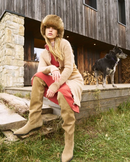 Adult female model wearing a fur hat, cardigan, dress, and boots, poses outside a wooden house next to a dog and woodpile. 