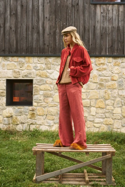 Woman in orange corduroy suit and cap posing outdoors on wooden pallet, stone wall, and grass.