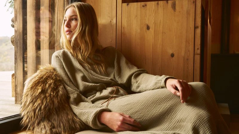 Adult woman relaxes in a light-filled rustic room. She is wearing a gauzy sage green outfit and seated next to a fur pillow.