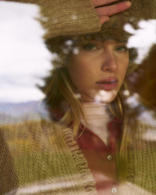 Portrait of a woman with light brown hair looking through a blurry window wearing a tan and white sweater, fur hat and red button shirt.