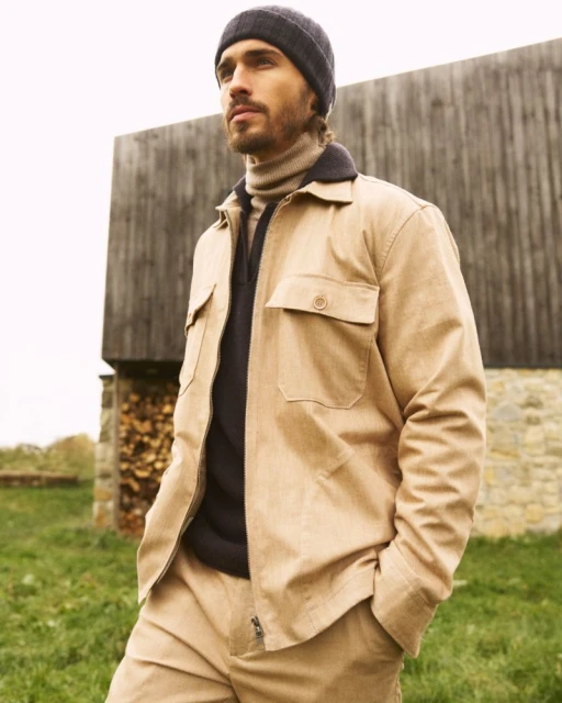 Adult man outdoors wearing a beige chore coat, dark beanie, and matching trousers, standing against a weathered wood and stone structure.