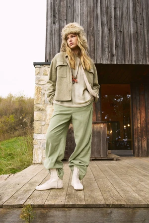Model wearing a fuzzy fur hat, green jacket, matching trousers, and white boots standing on a wooden deck.