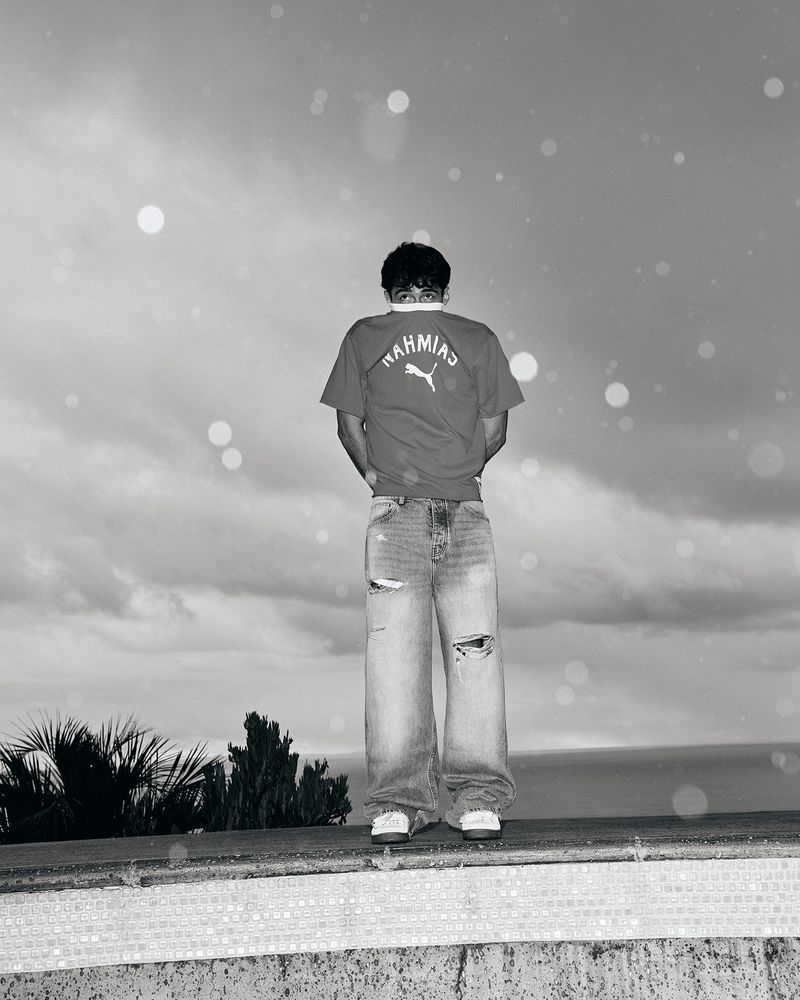 A person stands facing away wearing a red t-shirt and baggy jeans against a cloudy sky and ocean backdrop. A fashion or lifestyle image.