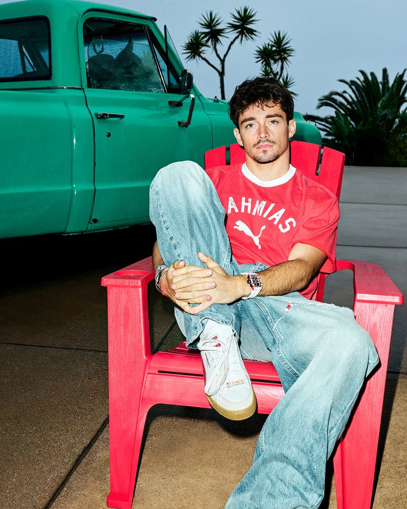 Charles Leclerc sits in an outdoor chair near a vintage turquoise truck and palm trees, wearing jeans, sneakers, and a red Puma t-shirt.