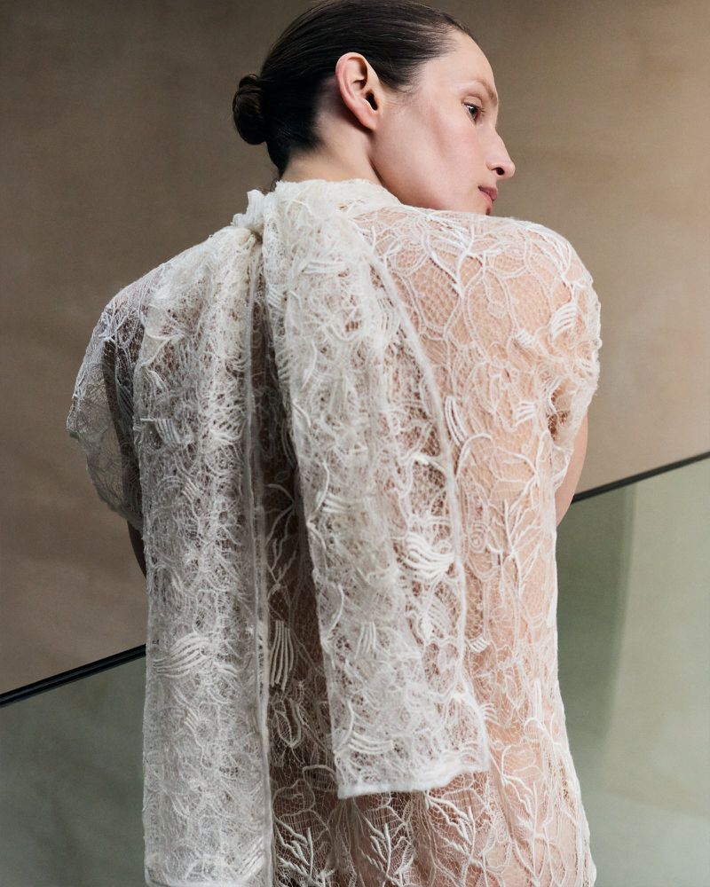 Elegant woman in a delicate white lace top, gazing away with a bun hairstyle against a soft, neutral-toned background.
