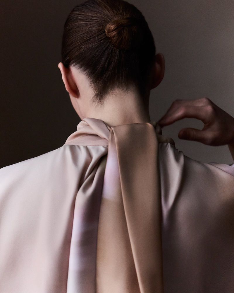 Elegant woman with a bun wearing a pale pink silky top, standing indoors with a neutral background, showcasing fashion and style.