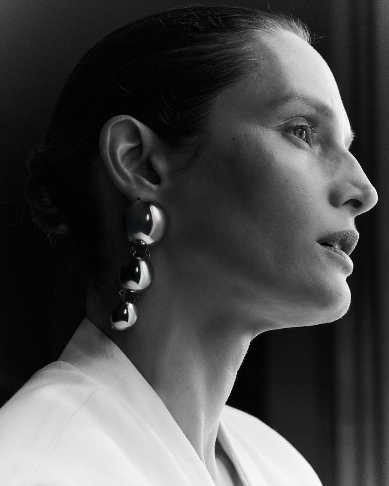 Close-up profile portrait of a female model wearing large metallic drop earrings and a white top against a dark background