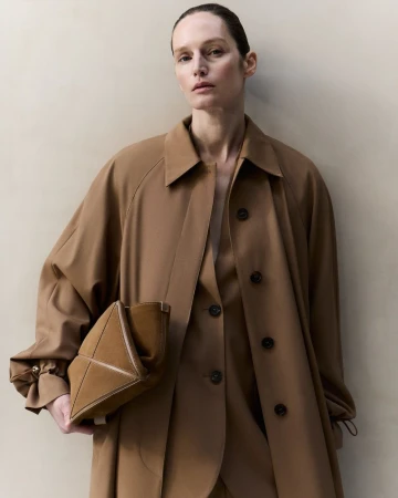 Fashionable adult female model poses in a studio wearing a tan overcoat, blazer, and slacks, holding a brown leather triangular handbag.