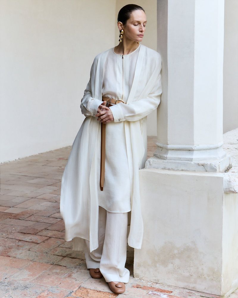 Elegant woman in an off-white linen suit with gold earrings stands in a covered hallway with terracotta tile flooring.