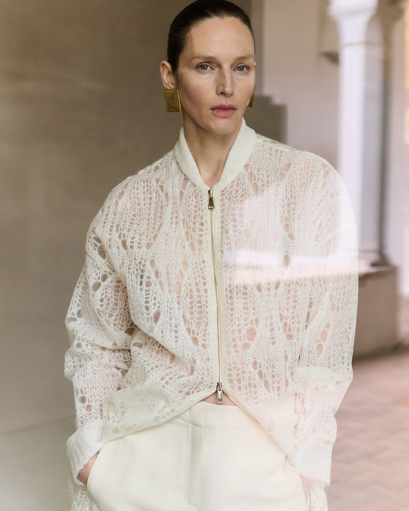 Woman in an airy, cream-colored, long-sleeved lace jacket and matching trousers, standing indoors with neutral wall and pillar background.