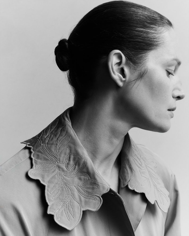 Monochrome portrait of a woman in a pale jacket with a decorative scalloped collar, styled with her hair in a bun and eyes closed.