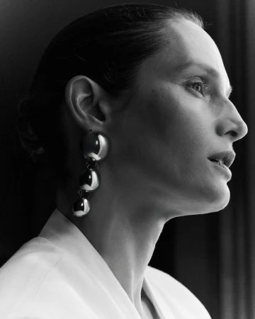 Close-up profile portrait of a female model wearing large metallic drop earrings and a white top against a dark background