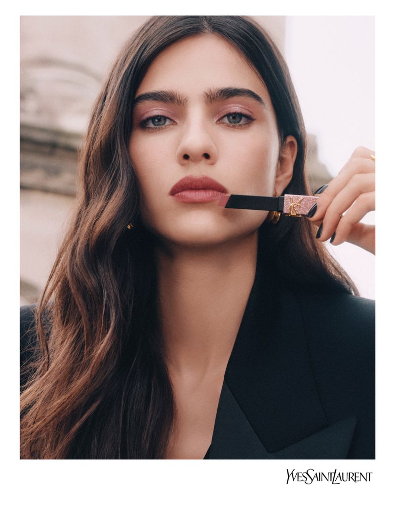 Close-up of a woman applying lipstick, showcasing the beauty product with natural makeup and styled hair, against a soft background.