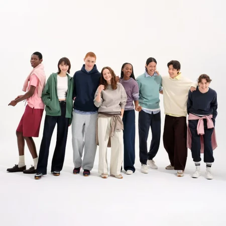 Group of eight diverse young adult models in casual clothing stand in a line against a bright white background in a fashion setting.