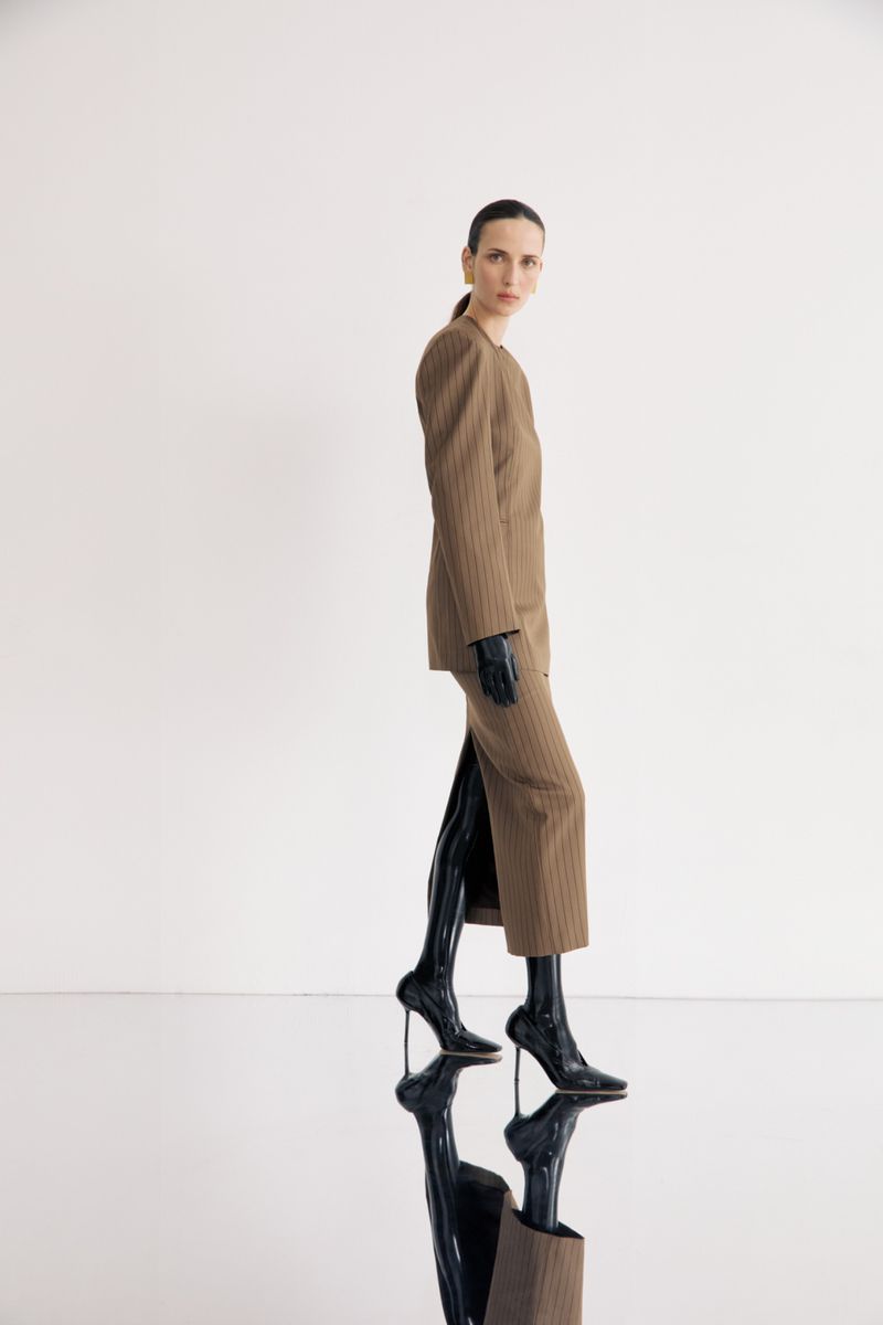 Woman in pinstripe suit poses in studio, glossy black boots, profile view, well-lit, clean background; fashion, style, portrait.