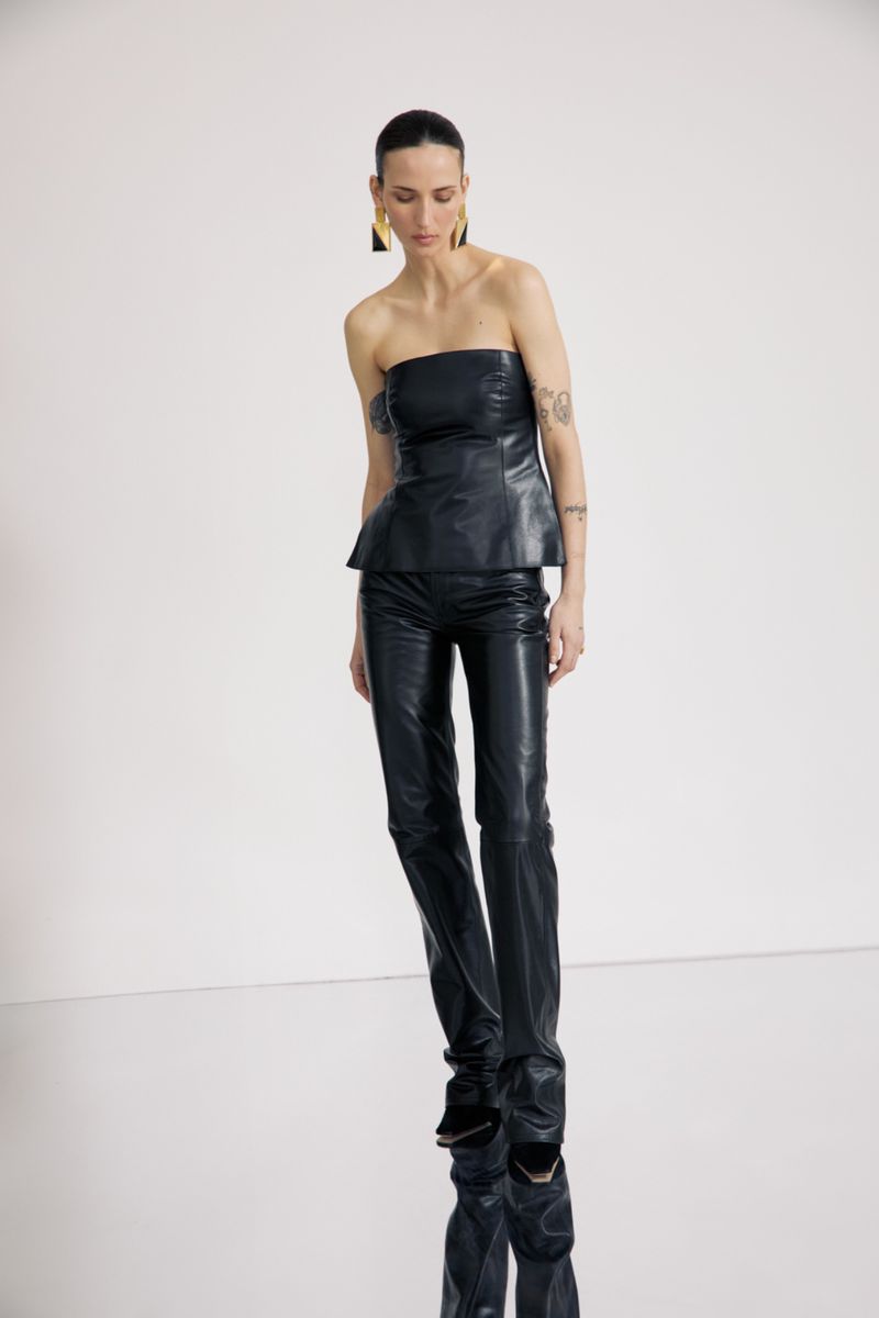 A slender woman in a dark, strapless leather top and matching pants poses against a clean white backdrop. Statement earrings add a modern touch.