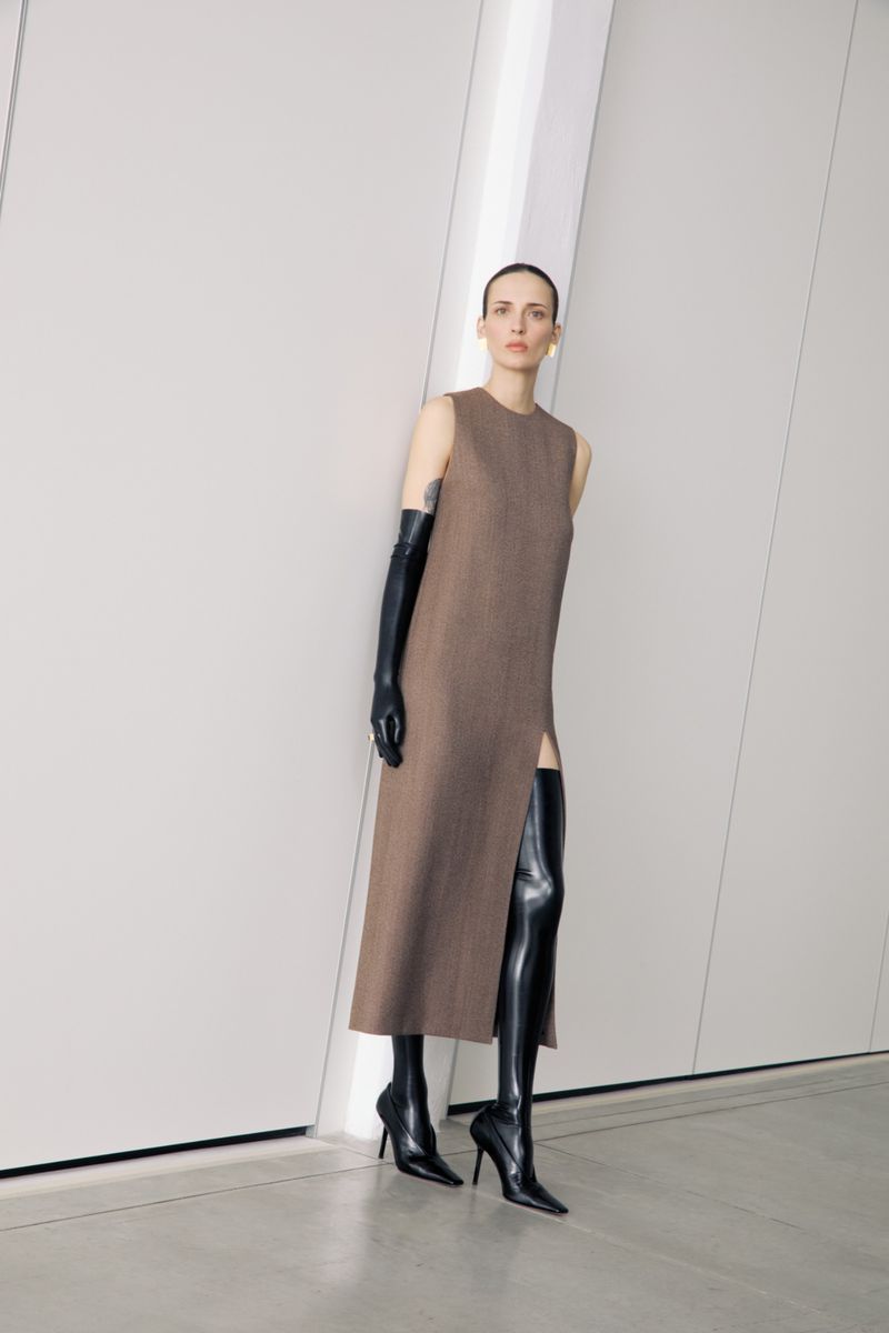 A woman poses in a sleeveless brown dress with a thigh slit, knee-high black boots, and black gloves against a white wall with soft lighting.
