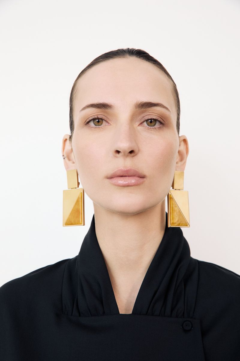 Headshot of a woman with dark hair pulled back, wearing large geometric gold earrings and a black top against a white backdrop.