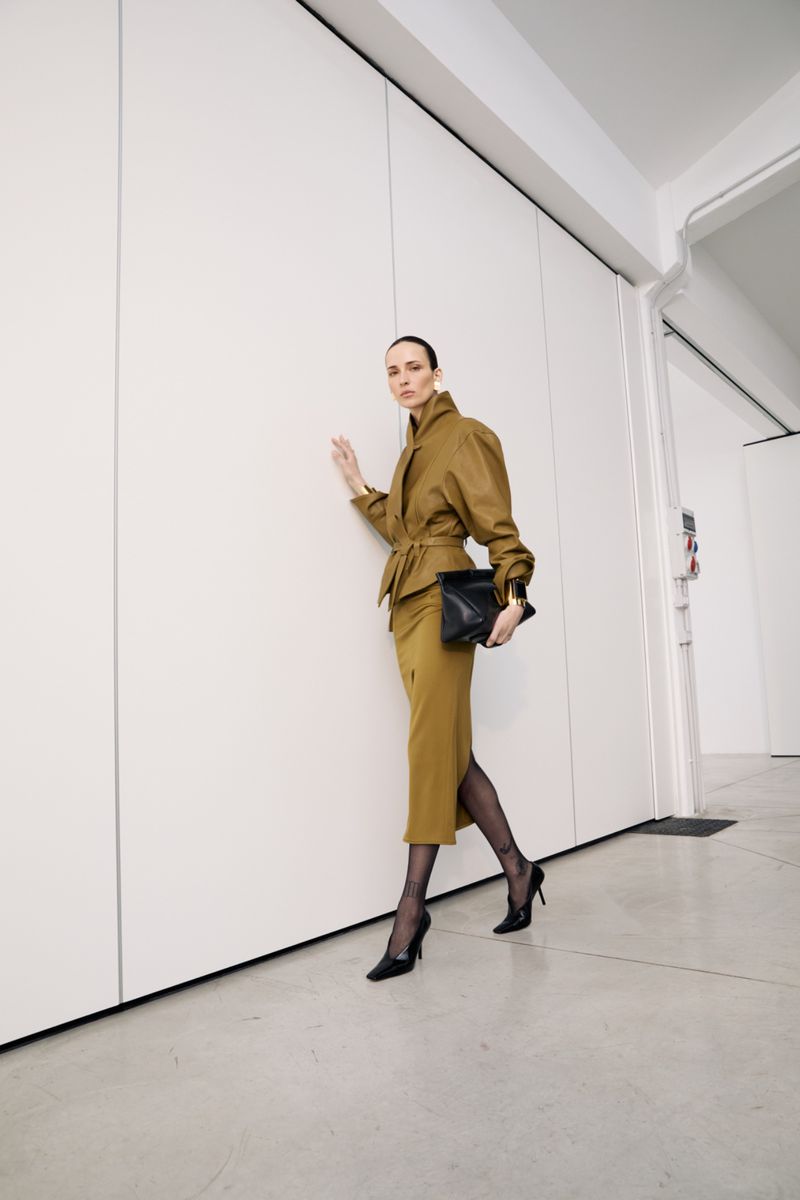 A woman in a golden-brown skirt suit poses in front of a white wall, holding a black clutch. Her outfit is completed with sheer black tights and heels.