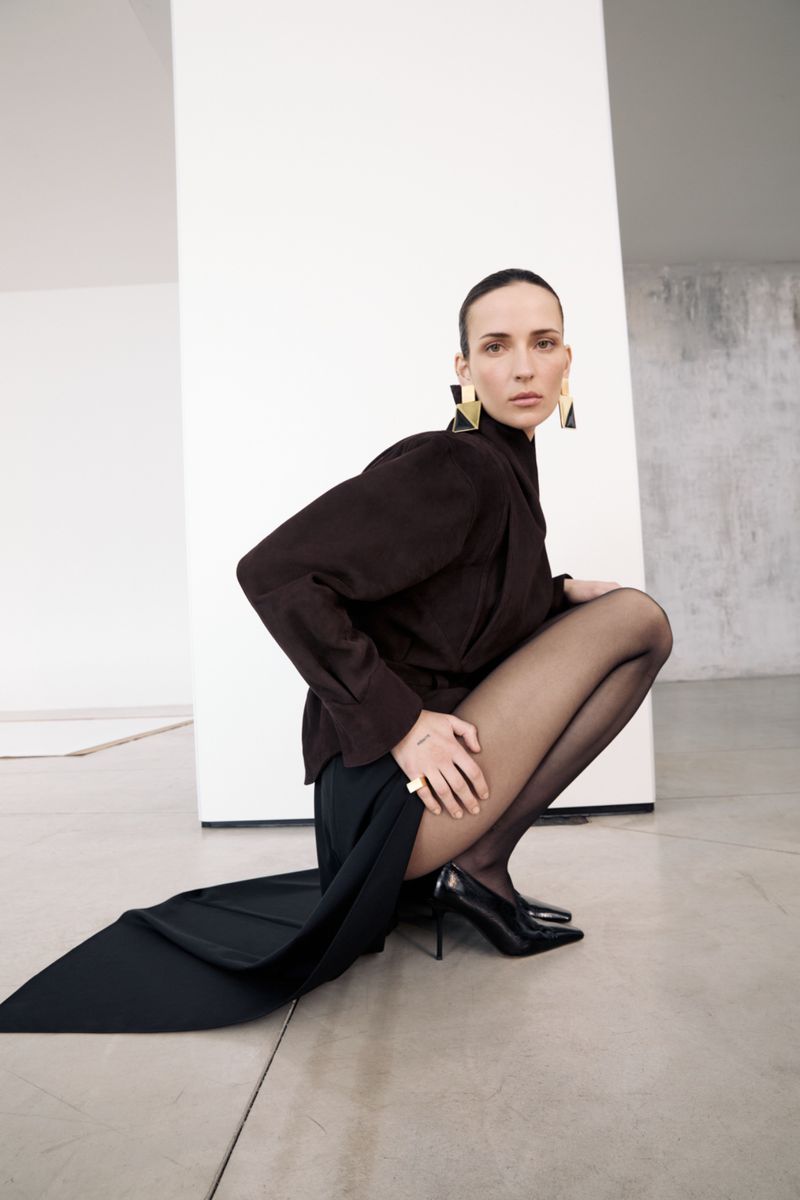 Brunette woman in a crouched pose wearing a dark jacket, skirt, black tights, and heels against a neutral studio backdrop with geometric shapes.