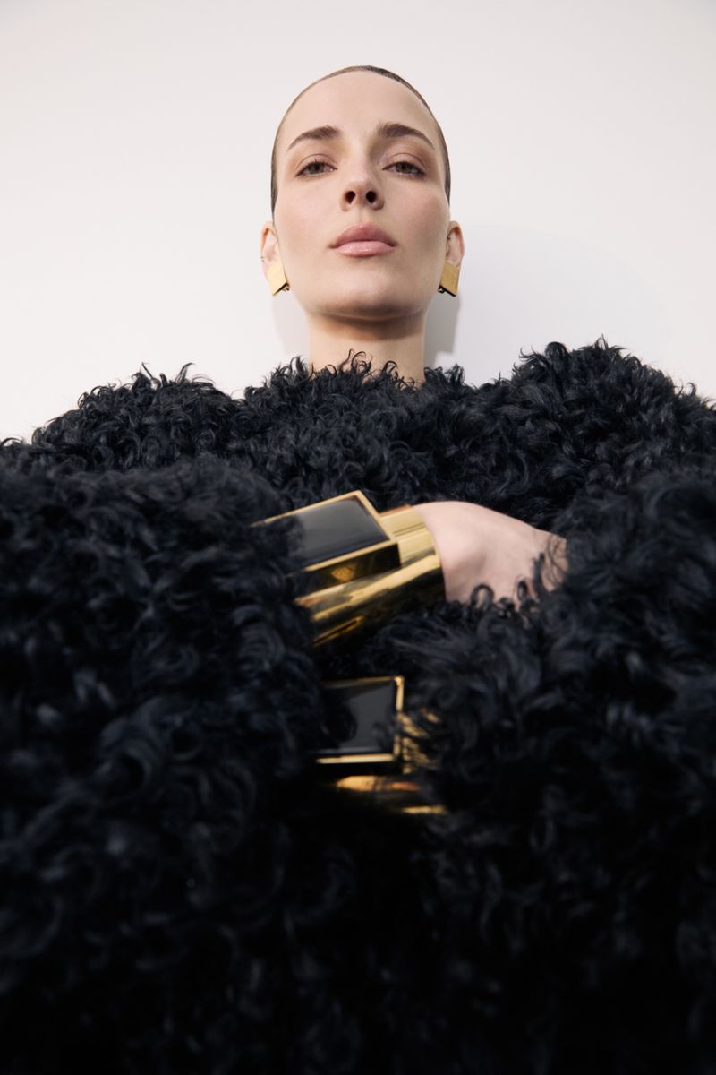 Woman wearing a black, fluffy jacket with large gold square bracelets and earrings, shot in a photo studio with a white background.