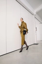 Model wearing a structured olive green jacket and skirt set with black heels and a black handbag against a white wall.