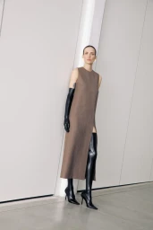 Model wearing a brown sleeveless dress with high black leather boots against a white studio wall