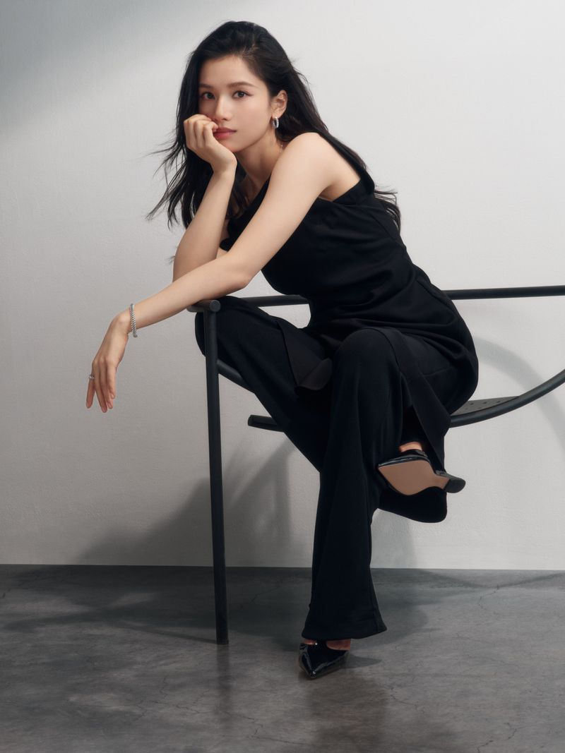 Portrait of a young Asian woman in a black outfit sitting on a black chair in front of a simple grey background.