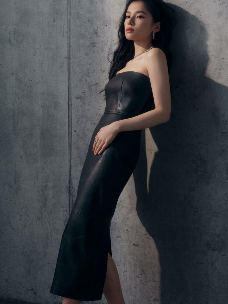 Portrait of a stylish adult woman with long dark hair wearing a strapless black dress and jewellery, leaning against a textured grey wall.