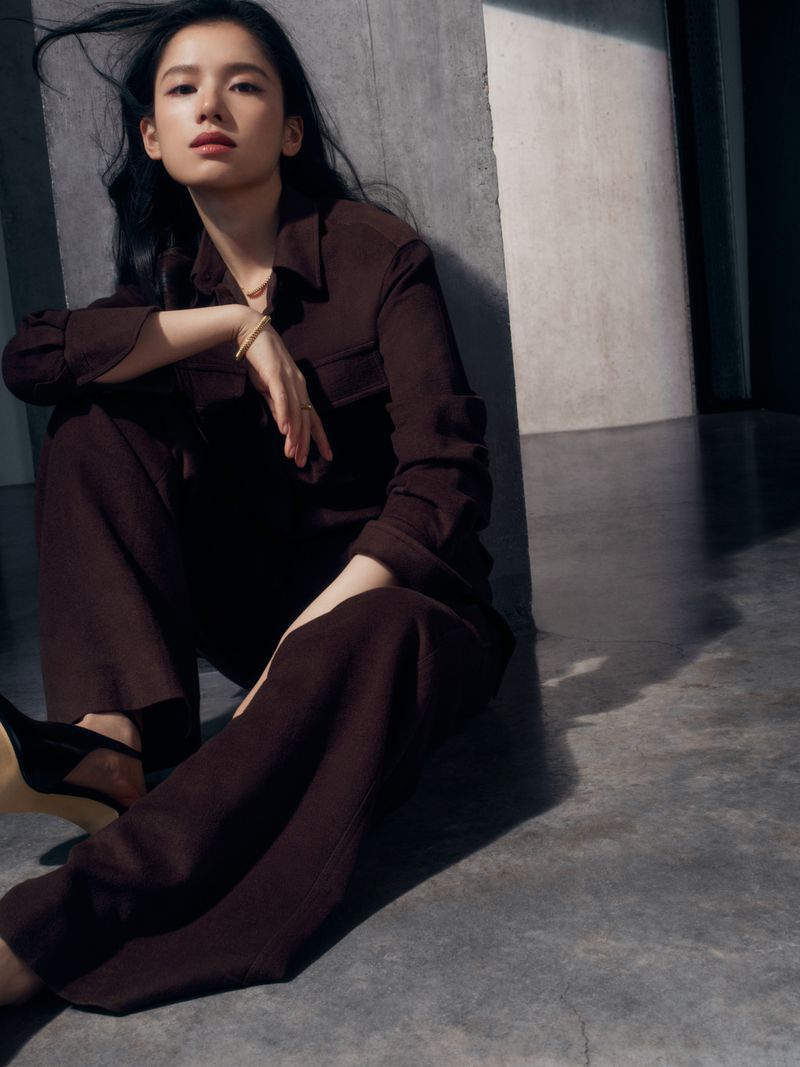 Portrait of a woman in a brown pantsuit sitting against a concrete wall, her dark hair cascading down her shoulders in soft waves.