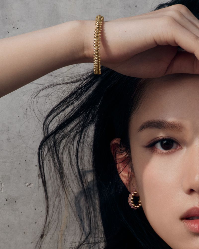 Close-up shot of a woman's face and arm wearing a golden bracelet and earrings against a textured gray background in soft lighting.