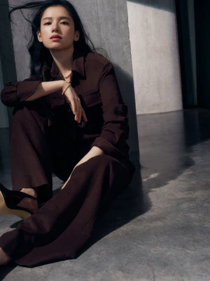 Zhang Jingyi in a brown button-down shirt and matching trousers sitting against a grey concrete wall for Cartier