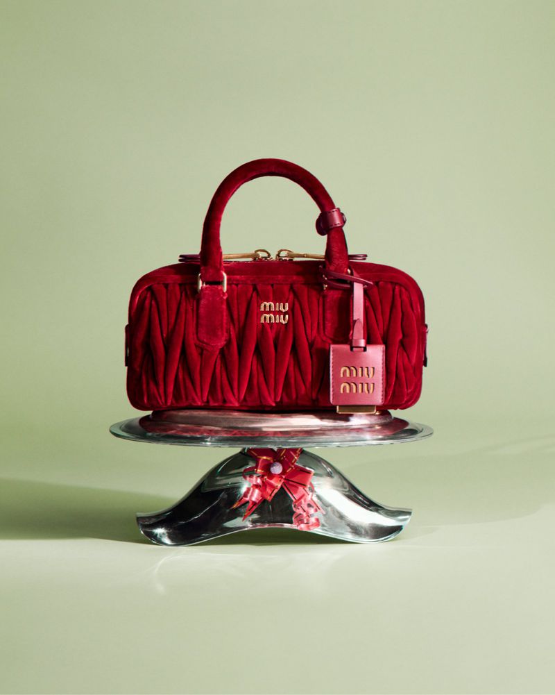 Red Miu Miu handbag with gold logo and tag, displayed on a silver cake stand against a pale green background in a studio setting.