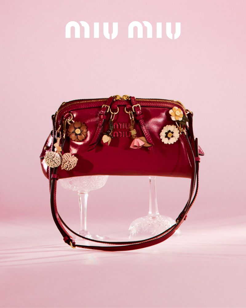 Red patent leather Miu Miu handbag with floral charms displayed on glass stands against a light pink background for a fashion editorial.