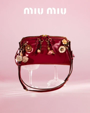 Red patent leather Miu Miu handbag with floral charms displayed on glass stands against a light pink background for a fashion editorial.