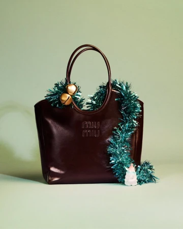 Dark brown Miu Miu leather tote bag with handles, adorned with blue tinsel garland and gold jingle bells against a pale green backdrop.
