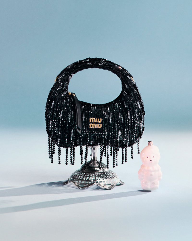 Miu Miu black beaded bag on a silver stand with a pink ornament on a light blue background in a studio setting.