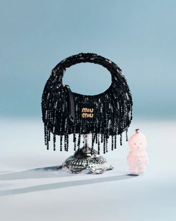 Black sequined Miu Miu handbag resting on a silver pedestal next to a small figurine against a light blue backdrop.