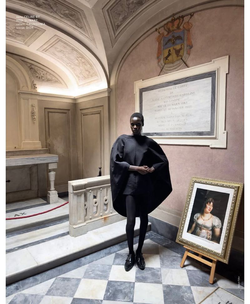 A solo female model in an all-black outfit poses inside a historic building with a checkered floor, a framed painting, and architectural details.