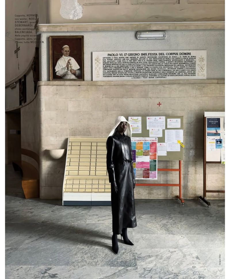 An African American woman in a black leather outfit and white head covering stands in a stark, tiled interior with religious imagery.
