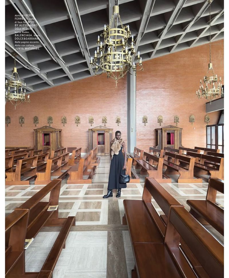Fashion shot in church interior: an African woman stands in a black dress, accessorized with a fur shawl and handbag in a church setting.