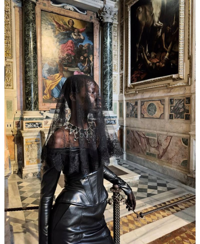 Woman in gothic fashion poses indoors near art. She wears a black leather dress, gloves, necklace, and lace veil in an indoor space.