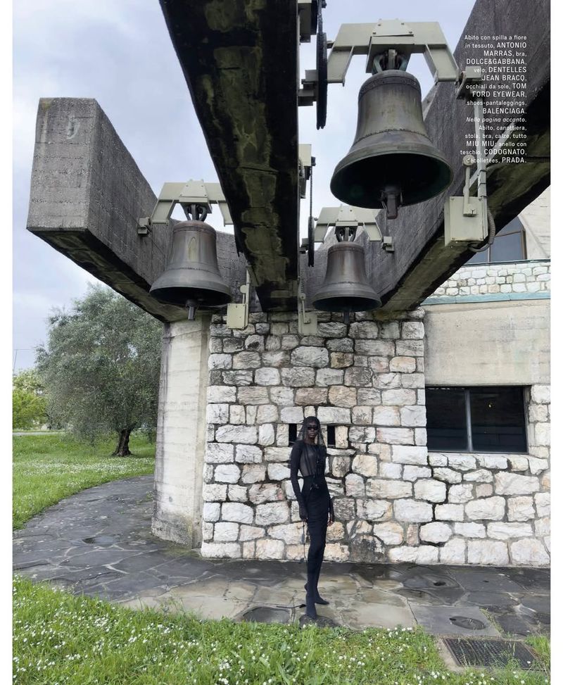 Fashion shot of a tall person in a black outfit standing under the bell tower of an old stone building in a grassy, outdoor setting.