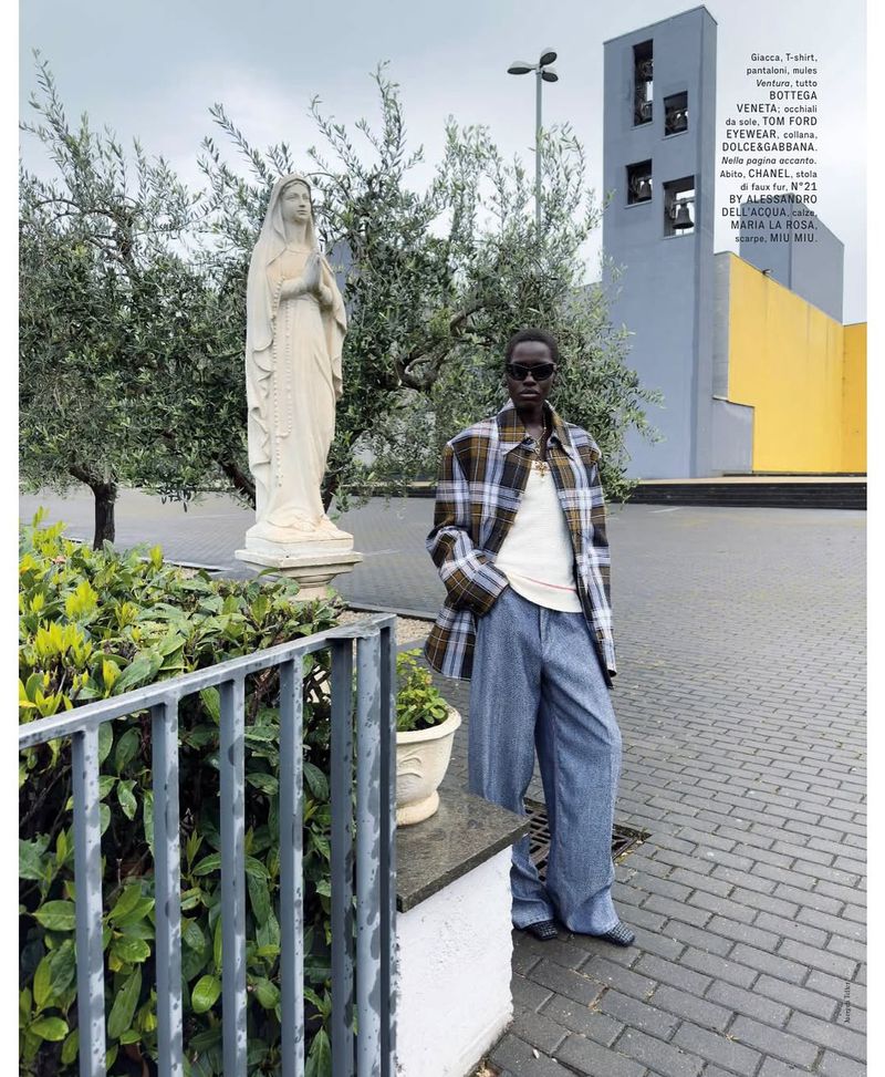 Fashion shot of a woman in a plaid jacket and jeans standing outside a church, with a statue of the Virgin Mary in the background.