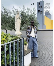 Model standing outdoors wearing a plaid jacket and blue trousers near a white statue and modern building.
