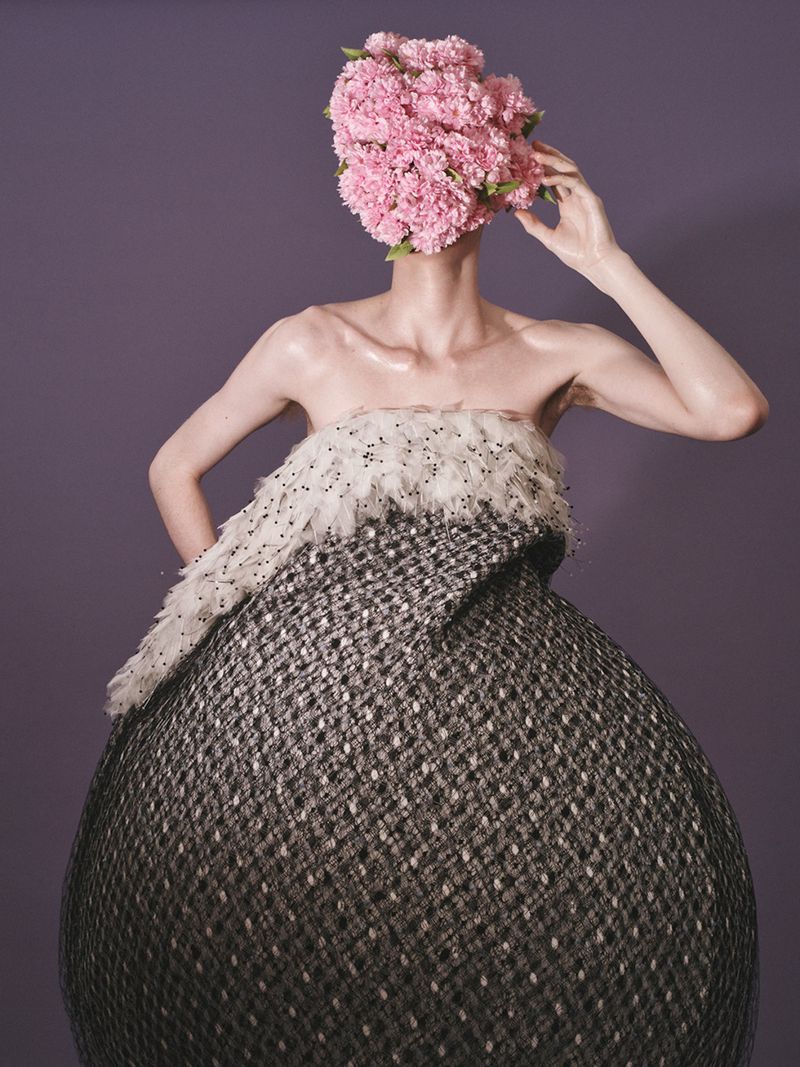 A person with a pink floral head covering wears a textured black and white gown against a solid purple background in a studio shot.