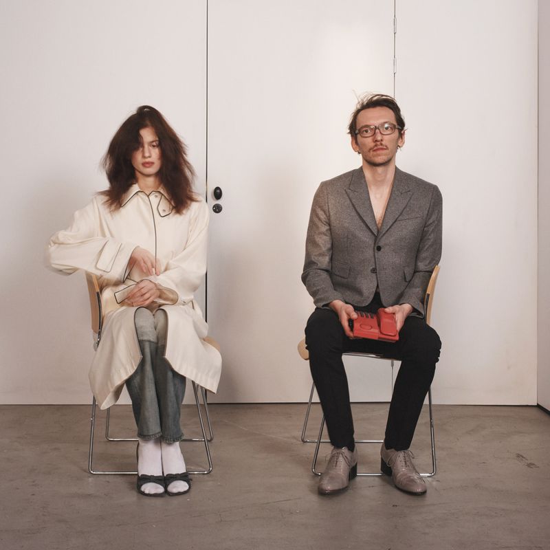 A seated adult woman in a white coat and an adult man in a grey jacket pose against a white wall in an indoor setting.